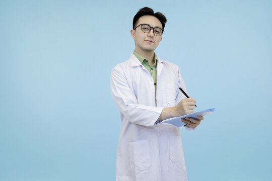 Confident Asian scientist or doctor in a lab coat and glasses, standing and  writing notes on a clipboard in blue studio background