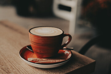 A warm cup of coffee with latte art sits on a wooden table in a cafe. The coffee cup is a vibrant orange color, and the latte art is a beautiful swirl. A sugar packet and spoon rest on the saucer.