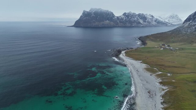 Aerial view of the rugged Uttakleivveien coastline, where turquoise waters meet the stark contrast of snow-capped mountains, Leknes, Nordland, Norway.