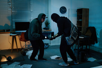 Two young adult men wearing masks and hoodies, breaking into office safe using flashlight,...