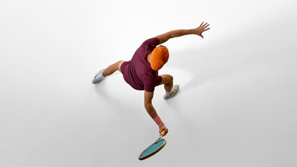 Male tennis player in orange cap reaching wide forehand from overhead angle on white floor. Concept of creative advertising, visual direction, storytelling movement, and athletic inspiration.