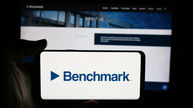 Stuttgart, Germany - 11-13-2025: Person holding cellphone with logo of US company Benchmark Electronics Inc. on screen in front of webpage. Focus on phone display.