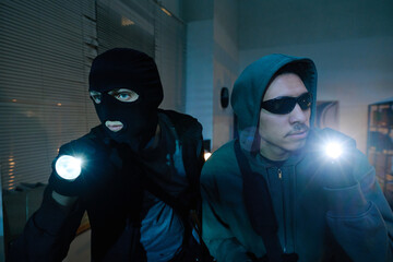 Two young adult men wearing masks and hooded sweatshirts, holding flashlights and sneaking through...