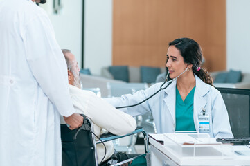 Old patient man sitting in wheelchair while young female doctor using stethoscope to check his heartbeat, elderly health checkup, happiness hospital.