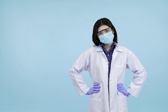 Confident Asian female scientist or doctor adjusting her lab coat and mask looking confidently in blue studio background