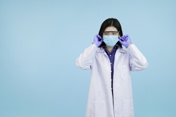 Confident Asian female scientist or doctor adjusting her lab coat and mask looking confidently in blue studio background