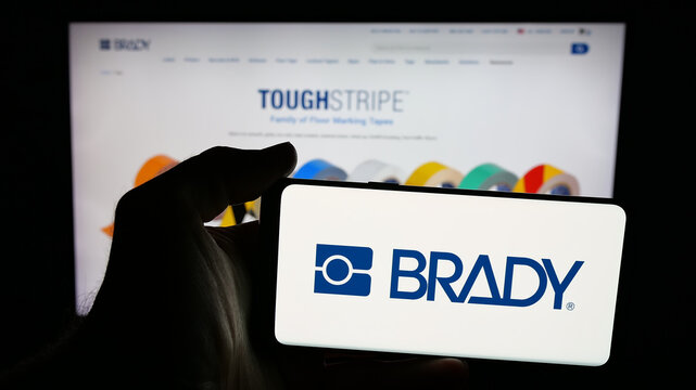 Stuttgart, Germany - 11-13-2025: Person holding mobile phone with logo of US manufacturing company Brady Corporation on screen in front of web page. Focus on phone display.