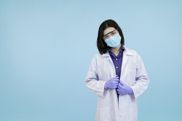 Confident Asian female scientist or doctor adjusting her lab coat and mask looking confidently in blue studio background