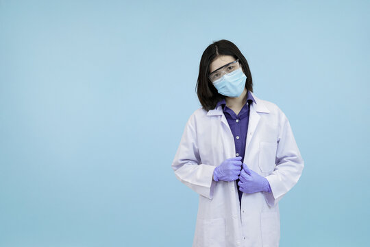 Confident Asian female scientist or doctor adjusting her lab coat and mask looking confidently in blue studio background - Powered by Adobe