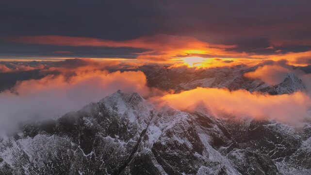 Aerial view of dramatic, snow-capped mountains piercing through a sea of clouds, illuminated by the warm glow of the setting sun, Ballstad, Nordland, Norway.
