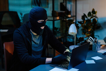 Young adult man wearing black balaclava sitting at desk using laptop in office environment, gloved...