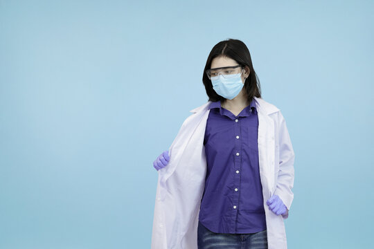 An Asian female scientist or doctor in a mask and safety glasses, adjusting her lab coat, symbolizing professional readiness and hygiene in blue studio background