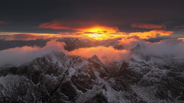 Aerial view of snow-dusted mountain peaks piercing through a blanket of clouds, illuminated by a fiery sunset, Ballstad, Nordland, Norway.