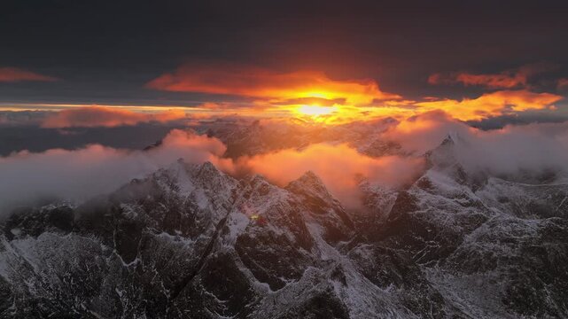 Aerial view of rugged, snow-dusted mountain peaks piercing through swirling clouds, bathed in the warm glow of the setting sun, Ballstad, Nordland, Norway.