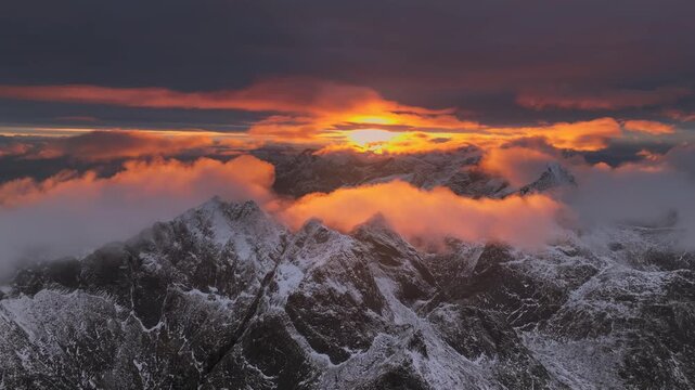 Aerial view of snow-dusted mountain peaks piercing through a layer of clouds, illuminated by the warm glow of the setting sun, Ballstad, Nordland, Norway.