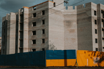 Construction of a new apartment building with a blue and yellow fence in front. The unfinished building stands behind a fence painted in the colors of the Ukrainian flag. Real estate market of Ukraine