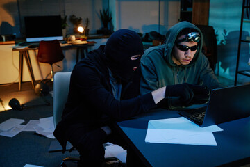 Two young adult men, one wearing balaclava and another one in hoodie and sunglasses, sitting at desk in office using laptop, stealing confidential information