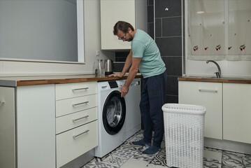 Man doing laundry at home in modern kitchen. Person loading washing machine and pouring detergent. Concept of household chores, domestic life, and home cleaning routine