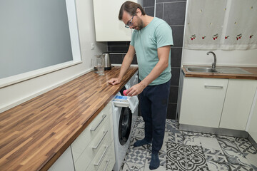 Man doing laundry at home in modern kitchen. Person loading washing machine and pouring detergent. Concept of household chores, domestic life, and home cleaning routine