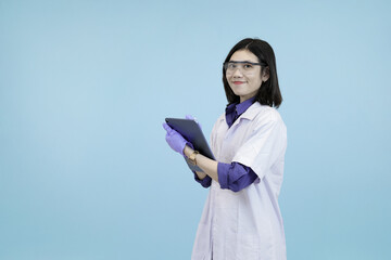 Smiling Asian female scientist or doctor in a lab coat and safety glasses, holding a tablet in studio blue background