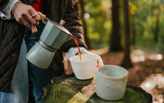 A woman is pouring coffee into two cups on a log. Scene is calm and peaceful, as the woman is enjoying a quiet moment in nature