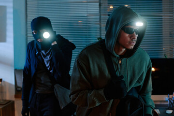 Two young adult men wearing masks and hoodies, sneaking through office at night, carrying bags and...