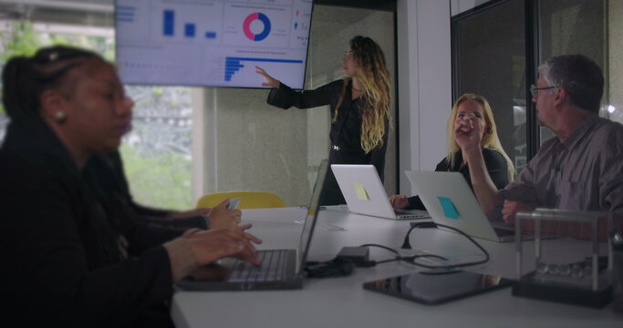 Confident woman stands in front of data screen as business team listens attentively during office meeting with laptops on table and focus on digital charts