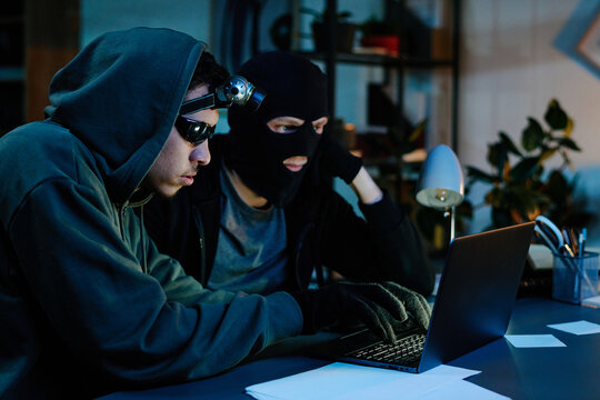 Two young adult men wearing hooded sweatshirts and masks, sitting at desk in office, using laptop while appearing to commit theft