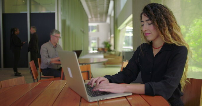 Confident woman typing on laptop at contemporary office lounge, side view showing concentration and serene workspace with nature outside large window - Powered by Adobe
