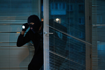 Man wearing black balaclava entering office at night holding flashlight, crouching near glass door,...