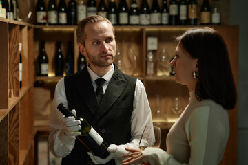 Caucasian middle aged man wearing formal attire holding wine bottle and presenting to Caucasian young adult woman holding wine glass in wine cellar, surrounded by shelves of bottles