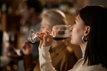 Young adult Caucasian woman tasting red wine in foreground, middle aged Caucasian woman and Black man sampling wine in background, people participating in wine tasting event