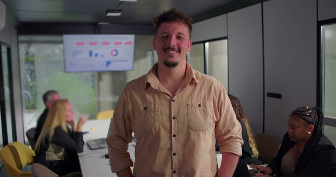 Confident young businessman with arms crossed in office, serious expression, diverse team focused on laptops in background near digital display
