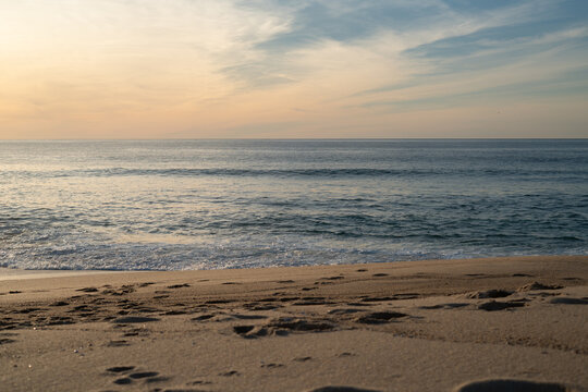 Early morning light over Lagoa de Albufeira beach. Footprints on sand, soft waves, and glowing sky.