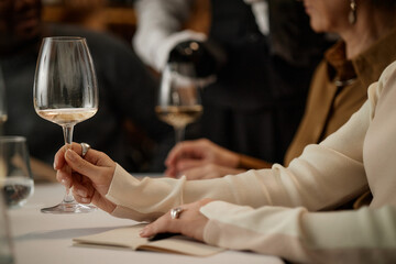 Caucasian young adult woman holding wine glass and swirling wine during tasting event, sitting at table with other adults, sommelier pouring wine in background, focus on hand