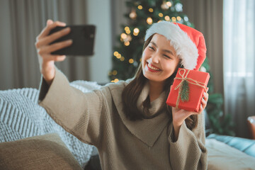 Joyful Asian woman taking a festive selfie with Christmas gift indoors, celebrating holiday season with warm lights, cozy winter vibes, cheerful lifestyle moment for Christmas and New Year