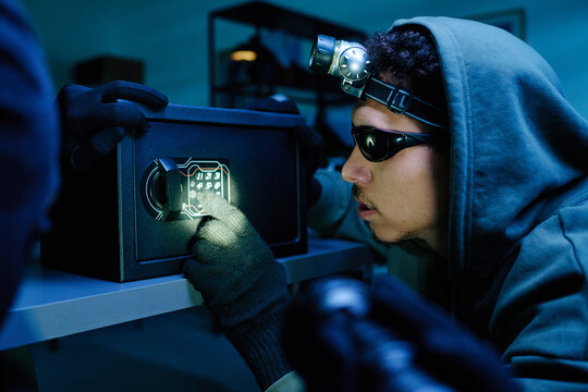 Young adult man wearing gloves and hoodie opening electronic safe in office, using flashlight attached to head, concentrating on keypad, committing theft during break-in