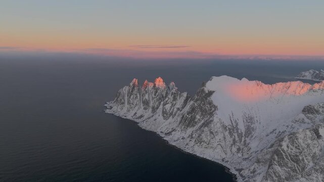 Aerial view of the rugged Okshornan peaks meeting the dark blue sea, their snow-covered tips kissed by the warm glow of the rising sun, Senjahopen, Troms, Norway.