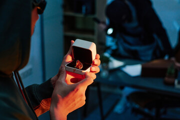 Young adult man holding jewelry box with large gemstone while middle aged man in background searching through office desk with flashlight during theft