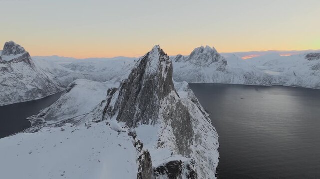 Aerial view of the snow-capped Segla mountain contrasting against the dark blue waters of the fjord under a pastel sky, Senjahopen, Troms, Norway.