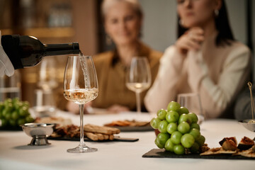 Two Caucasian women, one middle aged and one young adult, sitting at table tasting wine while sommelier pouring wine into glass, assorted snacks and grapes on table