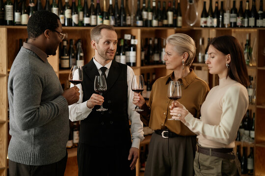 Group of middle aged Caucasian man, middle aged Caucasian woman, young adult Caucasian woman, and middle aged Black man holding wine glasses, standing together tasting wine in wine shop