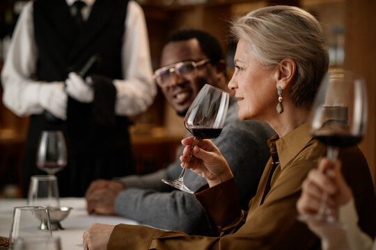 Middle aged Caucasian woman holding wine glass near face, smelling wine during tasting, sitting beside middle aged Black man smiling, sommelier standing in background serving wine