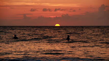 Sun dips below horizon as two surfers enjoy the tranquil ocean waves during a vibrant sunset at the beach, creating a peaceful and serene atmosphere.
