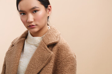 asian woman wears a taupe coat in a neutral studio setting, gazing softly at the camera. The calm portrait conveys elegance, poise, and mindful confidence.