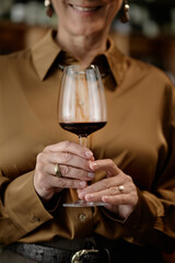 Cropped shot of middle aged Caucasian woman holding wine glass with both hands, smiling while preparing for wine tasting, focus on hands and glass, upper body partially visible