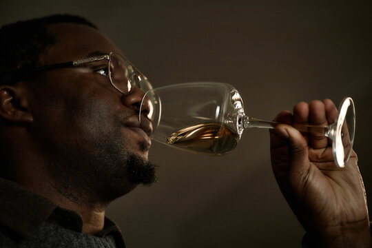 Black middle aged man holding wine glass near face, smelling and tasting wine during wine testing session, close up of profile with focus on sensory evaluation process