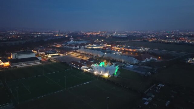Nighttime aerial view of the Ostra Dome in Dresden, brightly illuminated beside the sports fields and surrounded by the wide urban landscape.