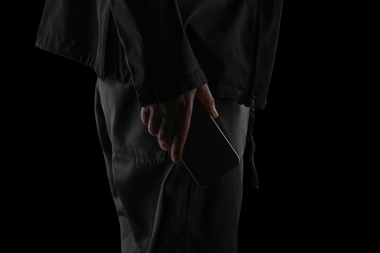 A close-up, low-key photograph of a person holding a blank smartphone beside their leg. Shot in a dramatic blackout aesthetic with deep shadows and soft highlights. Ideal for technology mockups, mobil