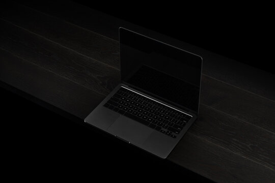 Photorealistic laptop mockup placed on a dark wooden surface in low-key lighting. Features a blank black screen, minimalist composition, and premium blackout aesthetic. Perfect for UI/UX showcases